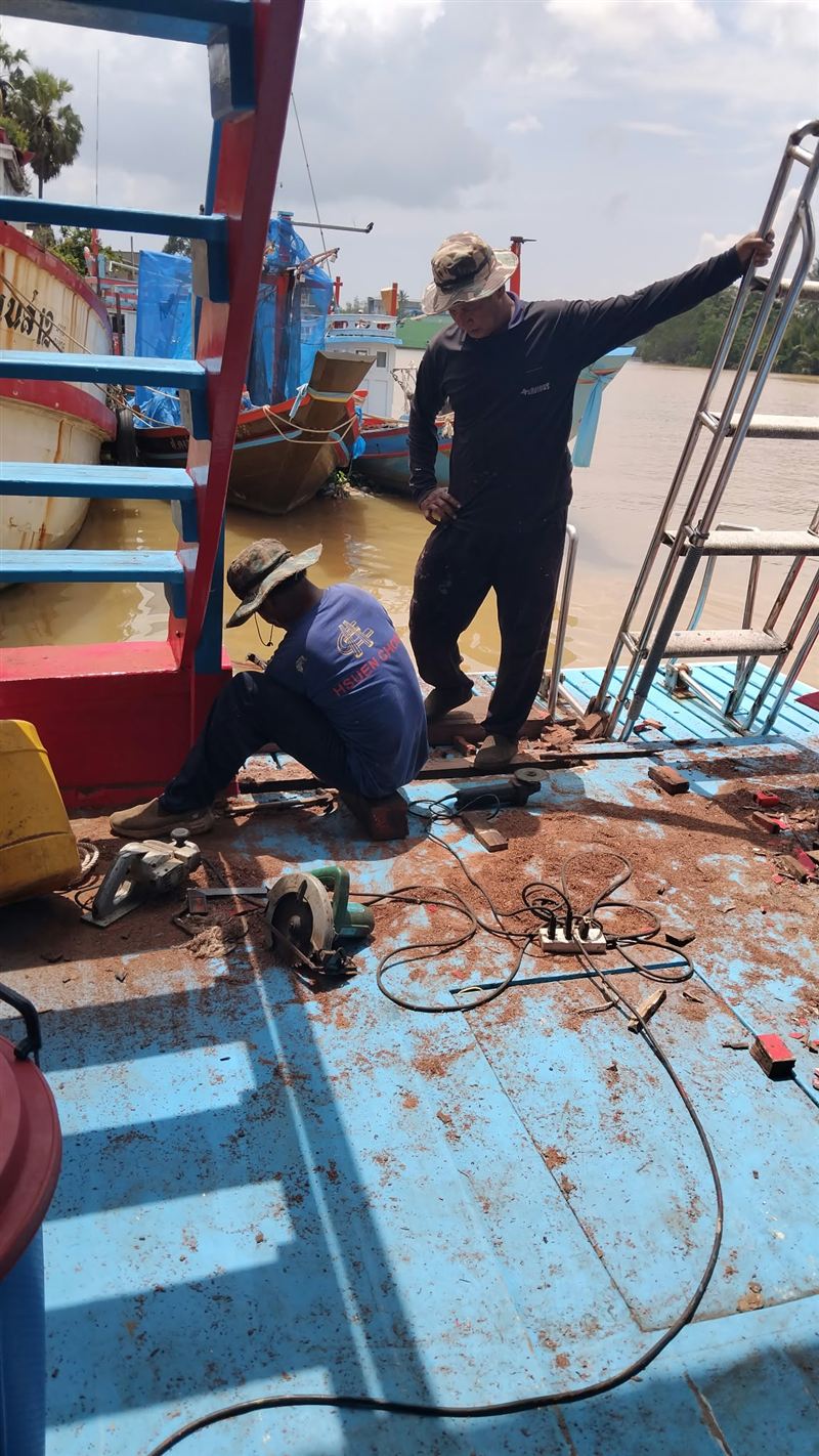 Workers fitting new parts on the back deck of the boat, with fishing boats on the river behind.